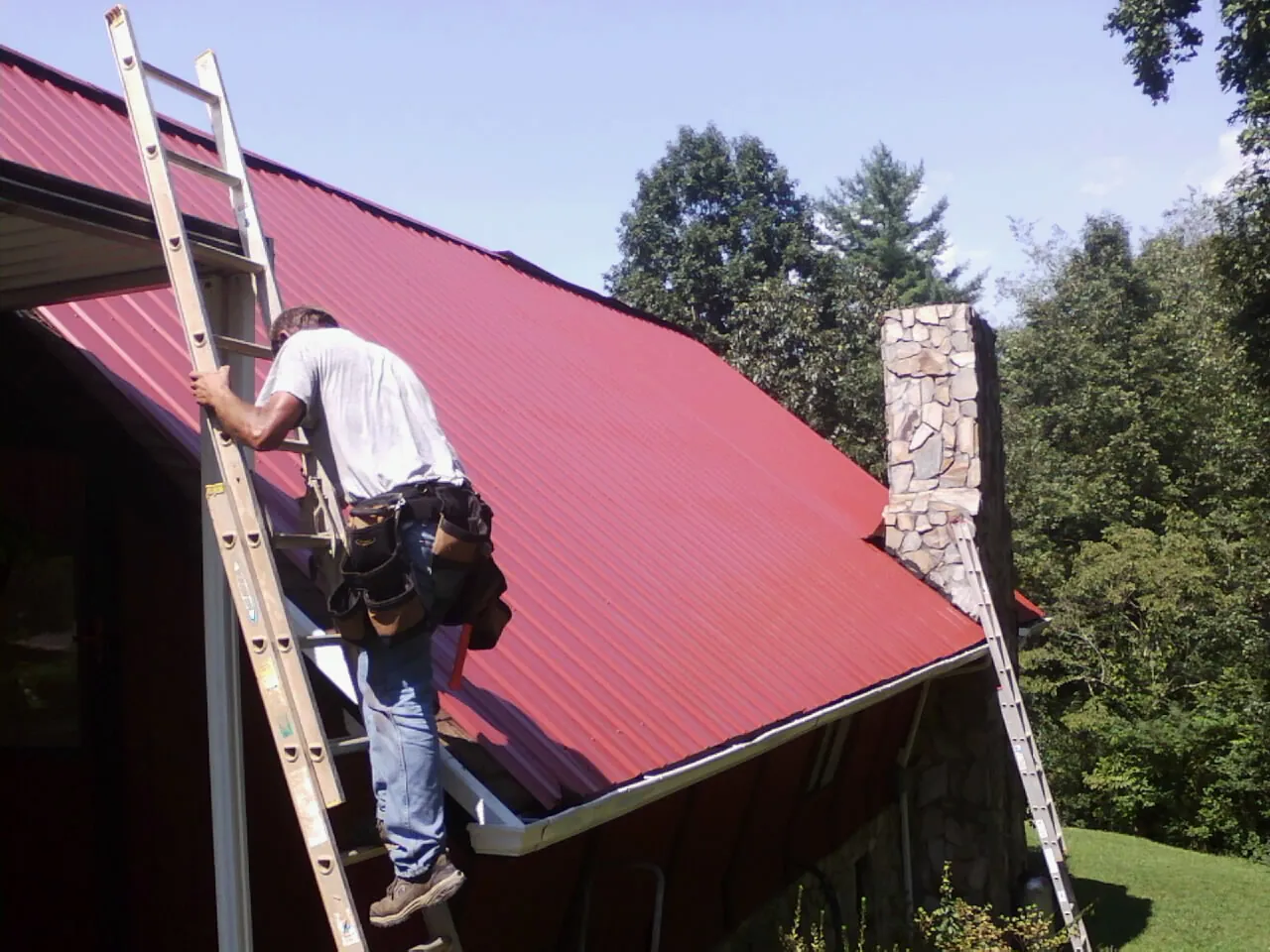 Roofer on a ladder beside a red metal roof and stone chimney — Soffit & Fascia Repair by Roofing Brothers in North Baltimore